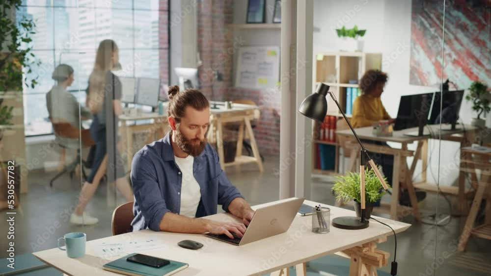 Poster handsome long-haired bearded manager sitting at a desk in creative office. young stylish man using l