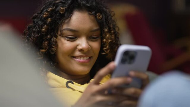 One happy African American young woman holding cellphone. A candid black Brazilian millennial girl looking at smartphone at night laid on sofa