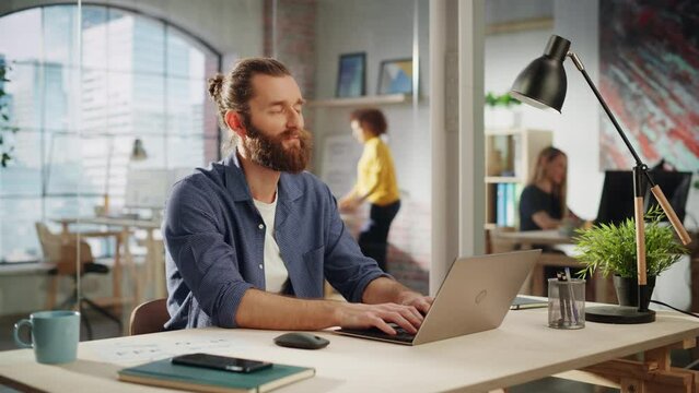 Creative Project Manager Sitting at a Desk, Working on a Laptop Computer in an Office. Boho Style Inspired Handsome Marketing Specialist Planning a Project, Browsing Internet and Writing Emails.