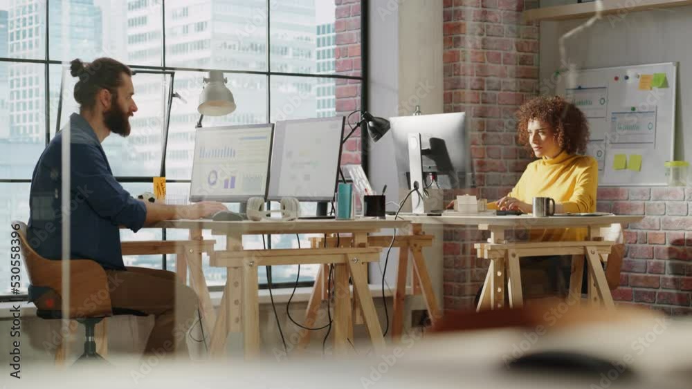 Poster Two Managers Sitting Behind Desks Next to Big Window with City View and Work with Analytical Reports on a Computer. Stylish Male and Multiethnic Female Working on Project in a Creative Office.