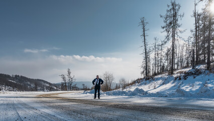 The highway curves along a snow-covered hillside. Snowdrifts on the roadside . There is a man standing on the road, a white scarf around his neck. Bare trees against a clear sky. Altai. Chuysky tract