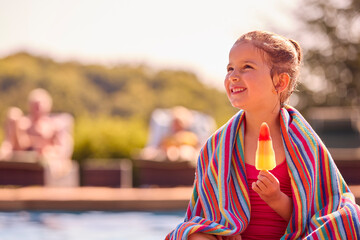 Family On Summer Holiday With Girl Wrapped In Towel Eating Ice Lolly By Swimming Pool