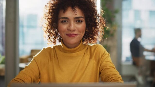 Portrait of a Beautiful Middle Eastern Manager Sitting at a Desk in Creative Office. Young Stylish Female with Curly Hair Looking at Camera with Big Smile. Colleagues Working in the Background.