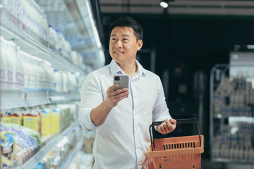 asian man shopper walks around the supermarket with a shopping cart looking at smart phone browsing...