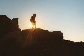 Naklejka premium silhouette of a woman with a backpack, against the light with the sun behind, hiking in the mountains. adventurous girl doing outdoor activity