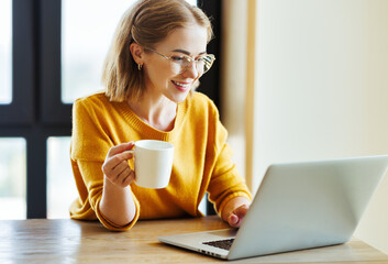 Cheerful young woman uses a laptop at home in kitchen with  cup of morning coffee - Powered by Adobe