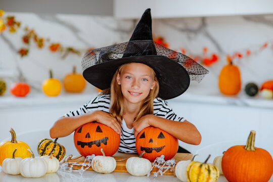 Girl in witch hat posing with coloured pumpkins for halloween on white kitchen.