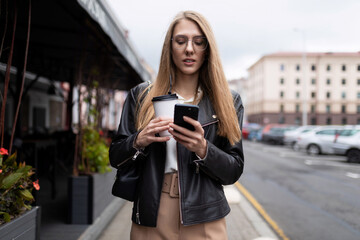 Fototapeta premium young woman typing a message on a mobile phone with a cup of coffee in her hands against the backdrop of the urban landscape next to a cafe