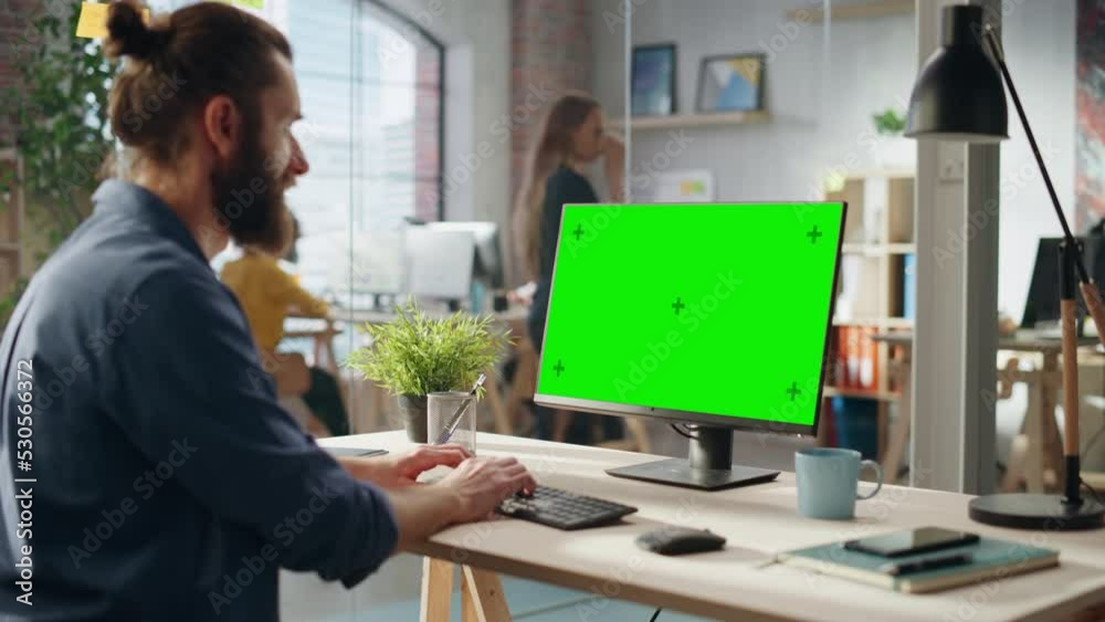 Poster Handsome Long-Haired Bearded Manager Working at a Desk in Creative Office, Using Desktop Computer with Green Screen Mock Up Display. Colleagues Working in the Background in Marketing Agency.