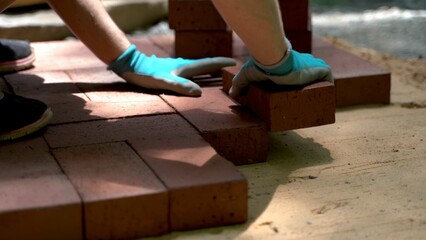 Low angle extreme closeup of person installing brick pavers onto sand base wearing blue gloves.