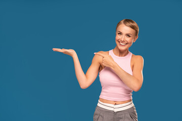 Smiling happy pretty young woman with short hair and natural makeup posing on blue studio background, pointing index finger aside.