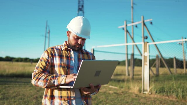 Design engineer in helmet holds laptop in hands at construction site in field, estimating investment attractiveness this object. Male compares his project with customer project. Using technologies.
