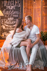 Obraz premium A young and attractive couple, a guy and a girl are sitting and hugging on a bed in a room decorated for Christmas with garlands. Christmas mood