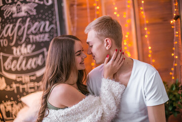 A young and attractive couple, a guy and a girl are sitting and hugging on a bed in a room decorated for Christmas with garlands. Christmas mood