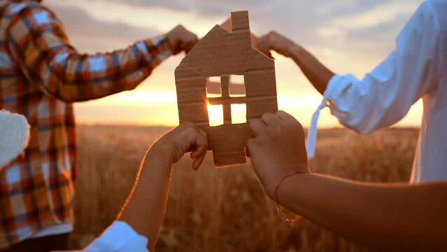 Close up small model paper house made of cardboard is held with hands family against on wheat field sunny sunset on summer nature. Concept home loans, mortgages, removal, ecology, building, economy.