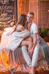A young and attractive couple, a guy and a girl are sitting and hugging on a bed in a room decorated for Christmas with garlands. Christmas mood