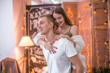 A young and attractive couple, a guy and a girl are sitting and hugging on a bed in a room decorated for Christmas with garlands. Christmas mood