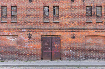 Wall of an old red brick factory building with narrow windows and metal gates