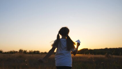 Follow to happy small girl running with a paper airplane through grass field. Cute child jogging among meadow and launching toy plane over sunset background. Concept of childhood dream. Rear view