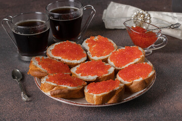 Homemade sandwiches with red caviar on white bread toasts on round plate and two cups of coffee on brown background