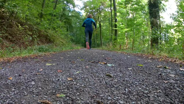 Jogger running on a gravel path in the forest filmed from behind. The shot starts as a close-up and ends in the distance.