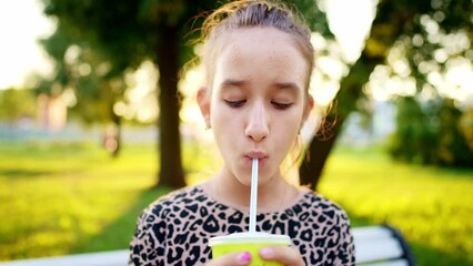 Young beautiful teenager girl sits in park on bench and drinks summer refreshing drink in plastic glass with straw. Delicious and healthy food. Leisure activity picnic in nature with green trees.