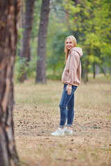Attractive blue eyed blonde woman walk on the city park. Girl wear beige hoodie, pink bag and look happy and smiles.