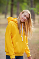 Blue-eyed blonde in a yellow hoodie walks in a pine forest. Portrait of a joyful young woman enjoying in autumn park.