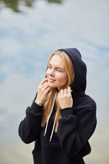 Beautiful young blonde woman in black hoodie walks in the park. Attractive female spending active...