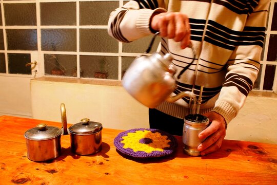Hombre cebando mate caliente a la ma&ntilde;ana con el desayuno sobre la mesa con la pava movida para dar movimiento, forma un original dise&ntilde;o de estilo de vida tradicional con fondo del ventanal con vidrios
