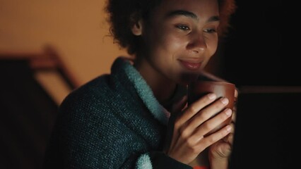 Positive African woman covered with a blanket looking at laptop with cup of tea in mountains