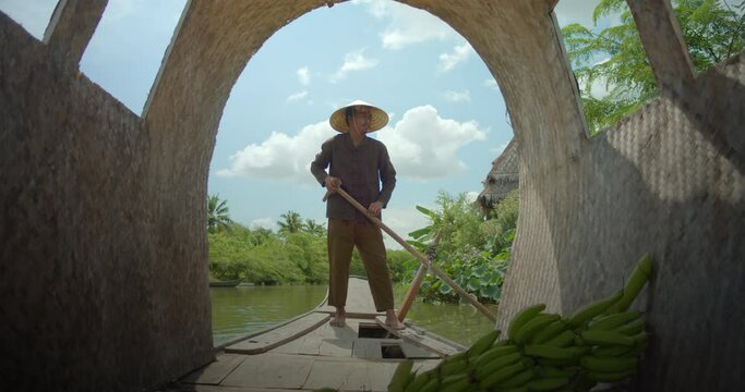 Slow motion an Asian male farmer who is native, stands and rows a boat in a canal that surrounds his banana plantation. And on holidays, he will take tourists on a cruise along the natural canal.