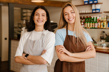 Two young waitress women wearing aprons smiling while working at cafe
