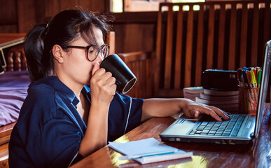 Young modern Asian woman in a casual outfit and eyeglasses sitting at a table with a notebook working on a laptop while having a mug of morning coffee at home