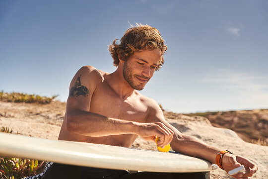 Caucasian Tattooed Surfer Cleaning His Surfboard And Removing Old Wax With Wax Comb