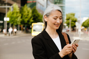 Grey asian woman smiling and using mobile phone at street