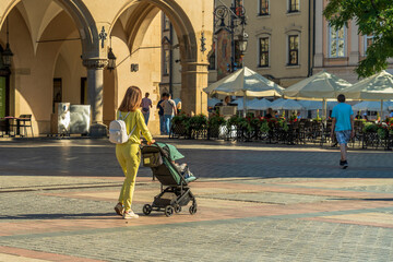 A young mother with a white backpack on her back walks with a baby stroller in the city square in...