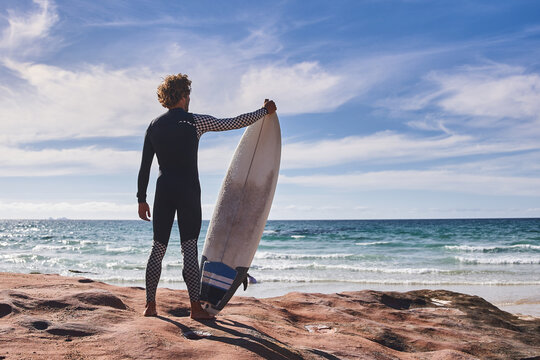 Back view of the man surfer carrying his surfboard and looking distance - Powered by Adobe