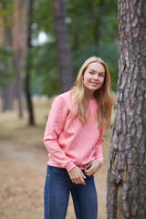 Obraz premium Blue-eyed blonde in a pink hoodie walks in a pine forest. Portrait of a joyful young woman enjoying in autumn park.