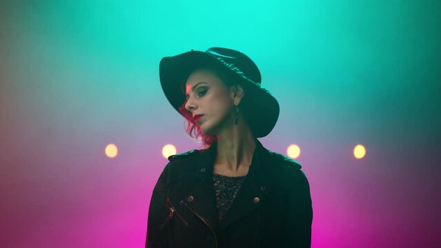 Female model, actress filming in a studio filled with smoke and neon lights background. Young woman in a hat looking around in the middle of the room.