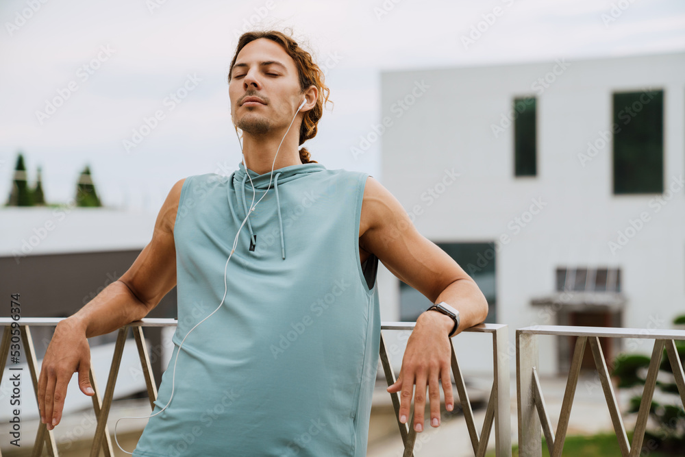 Wall mural Young athletic long-haired man in headphones relaxing with closed eyes