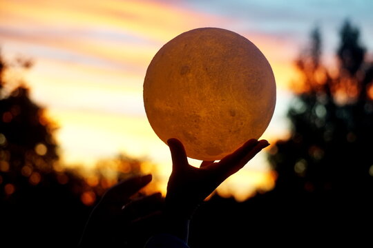 Moon In The Hand On Red Sunrise Background. Moon Bedside Lamp On The Sky