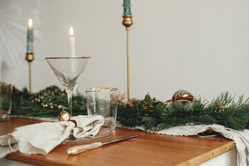 Stylish Christmas table setting. Linen napkin with bell on plate, vintage cutlery, wineglass, fir branches with golden lights, pine cones and candle on table. Atmospheric Holiday brunch