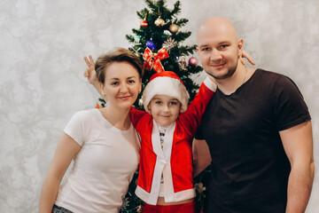 Family mom, dad and son in Santa costume are standing near the Christmas tree in their apartment and looking into the camera