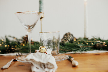 Stylish Christmas table setting. Linen napkin with bell on plate, vintage cutlery, wineglass, fir branches with golden lights, pine cones and candle on table. Atmospheric Holiday brunch