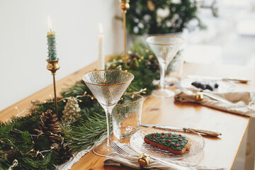 Stylish Christmas table setting. Christmas tree cookie on plate, linen napkin with bell, vintage cutlery, wineglass, fir branches with golden lights, candles on table. Atmospheric Holiday brunch