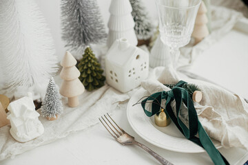 Christmas table setting. Stylish napkin with ribbon and bell on plate, vintage cutlery, modern festive christmas trees and houses on white rustic table. Holiday arrangement of table