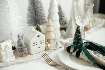 Christmas table setting. Stylish napkin with ribbon and bell on plate, vintage cutlery, modern festive christmas trees and houses on white rustic table. Holiday arrangement of table