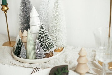 Christmas table decor. Stylish christmas little trees on plate, linen runner, golden candles on rustic table. Modern white artificial fir trees decoration in room. Holiday arrangement, table setting