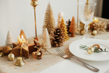 Christmas luxury table setting. Cedar branch with bell on plate, vintage cutlery, glasses, golden little christmas trees and ornaments on table. Holiday arrangement of table